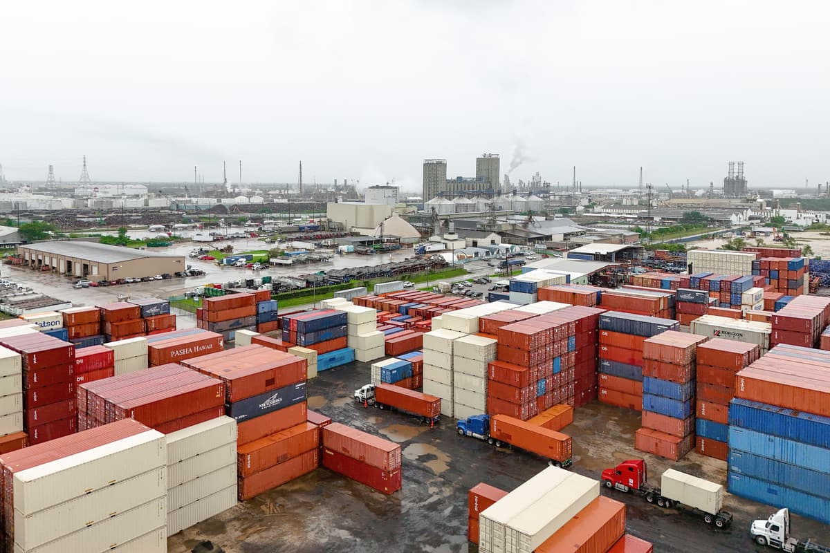Rows of stacked red, blue and white shipping containers in a storage yard in Galena Park, with trucks and industrial plants in the background