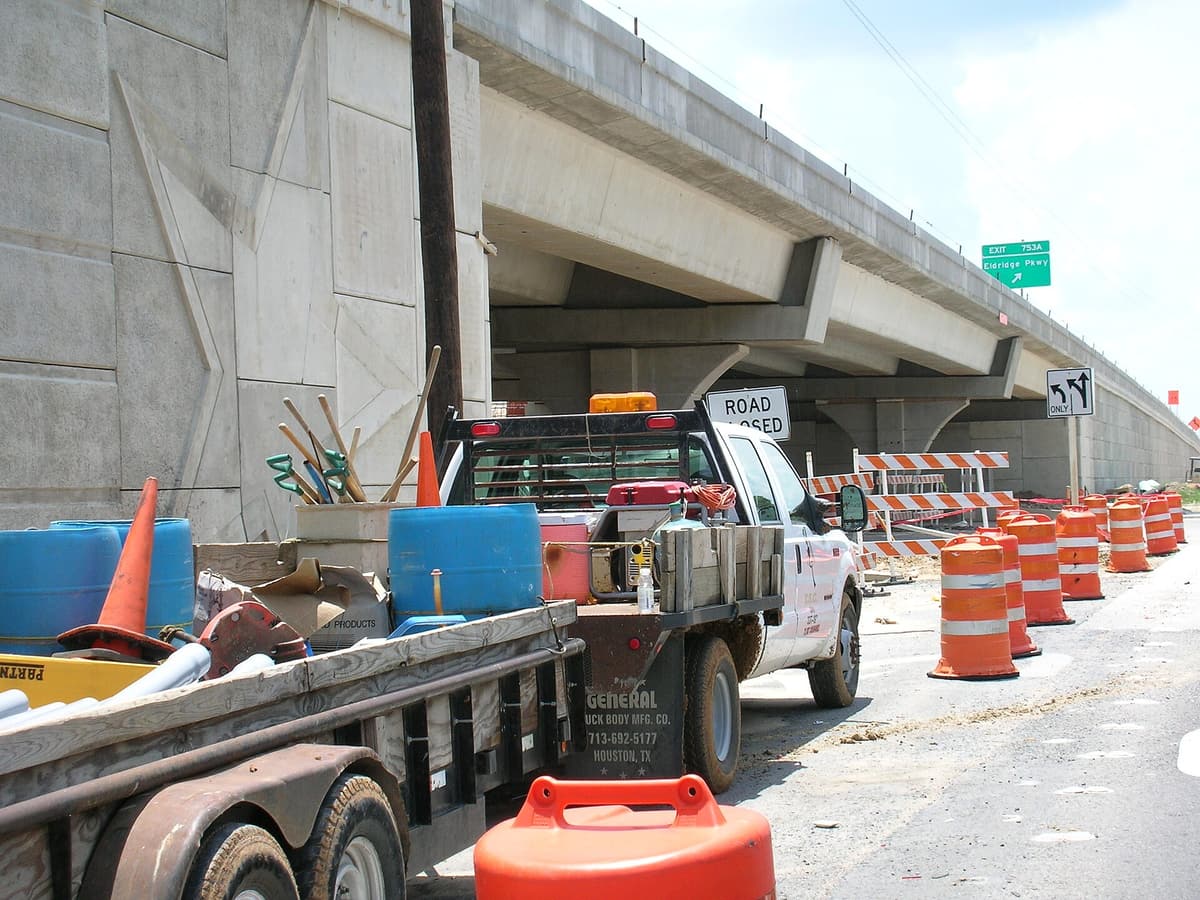 Orange traffic barrels and a Road Closed sign in an Interstate 10 work zone, with a contractor's pickup and trailer parked beneath a new concrete overpass
