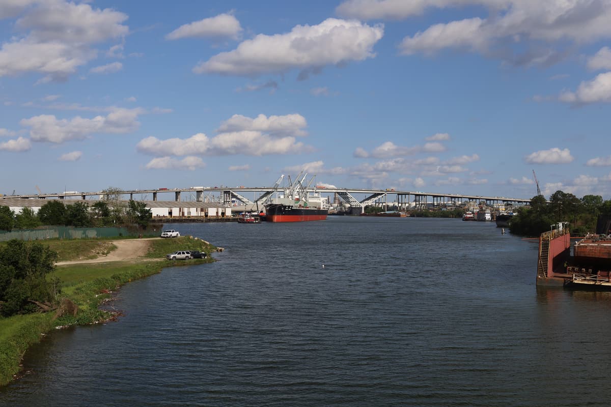 The Sidney Sherman Bridge spanning the Houston Ship Channel, with a bulk cargo ship and a tugboat passing beneath it under a partly cloudy sky