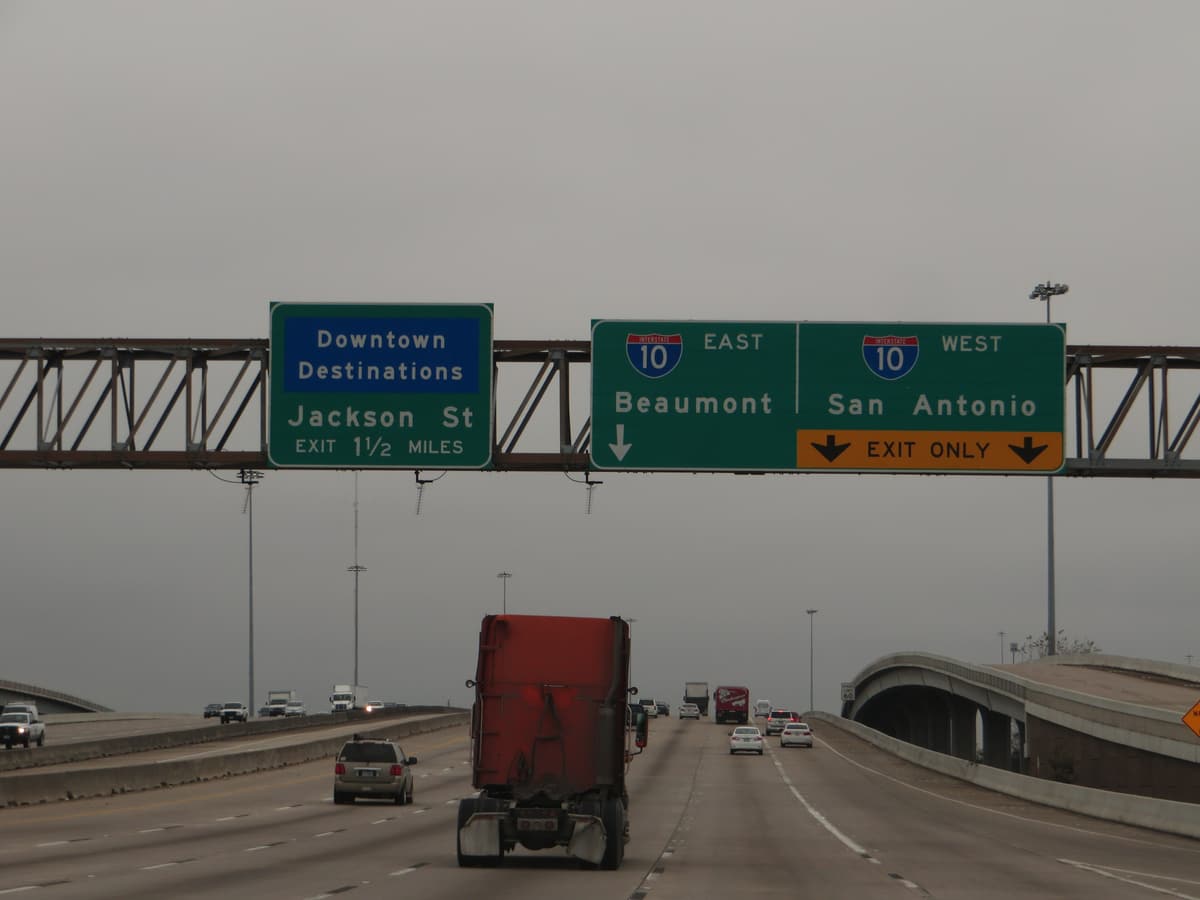 Overhead sign gantry on U.S. 59 in Houston with signs for downtown, I-10 East to Beaumont and I-10 West to San Antonio, above traffic that includes a red tractor-trailer